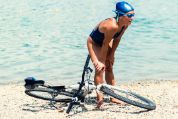 Eine Triathletin trägt einen Badeanzug, Bademütze und Schwimmbrille und hat nasse Haut. Sie stützt sich mit den Armen auf ihre Knie ab. Hinter ihr sieht man Wasser. Und bei ihr liegt ein Fahrrad auf dem Kieselstrand.  Eine Triathletin trägt einen Badeanzug, Bademütze und Schwimmbrille und hat nasse Haut. Sie stützt sich mit den Armen auf ihre Knie ab. Hinter ihr sieht man Wasser. Und bei ihr liegt ein Fahrrad auf dem Kieselstrand.